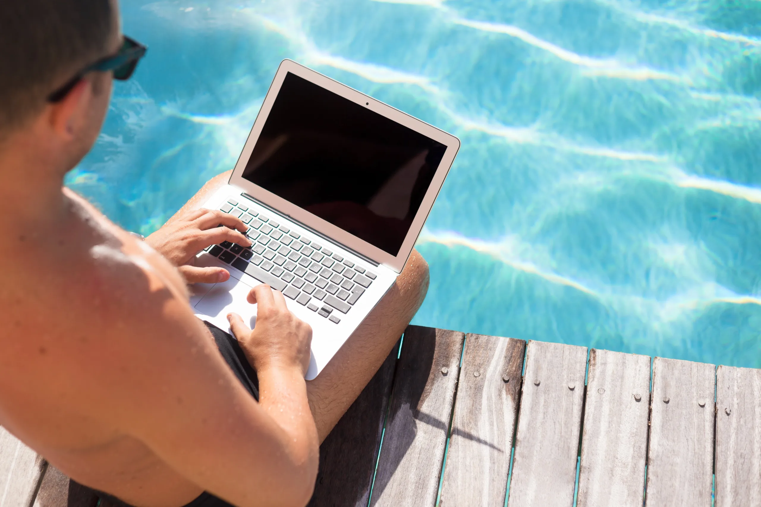 A man in a swimsuit by his crystal-clear pool in the sunshine using a laptop with sunglasses on. Photographed from over his right shoulder with space of viewing the pool and deck to the right of him.