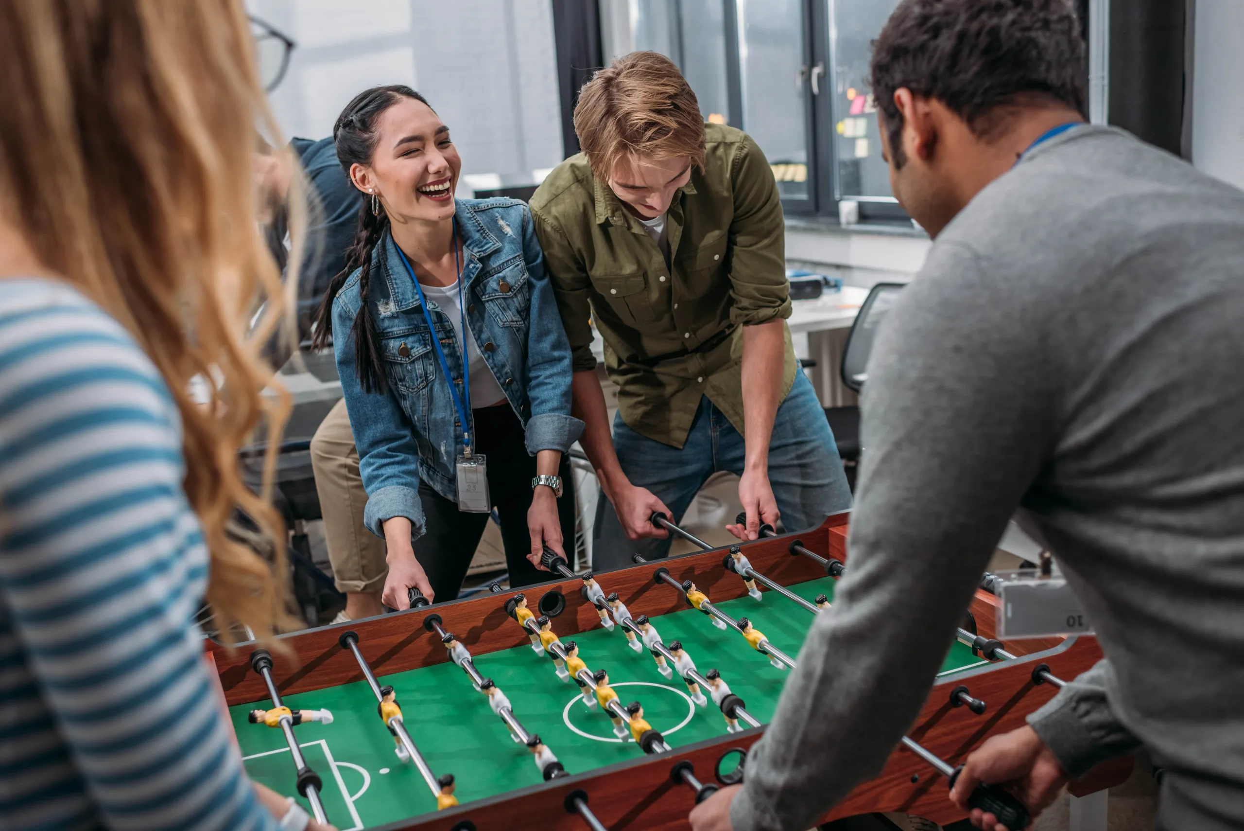 Men and women playing foosball in the game area of a clubhouse. One woman is laughing and the man beside her is looking down intrigued and focused on the game while another man and woman are in the foreground also participating from the other side of the table.