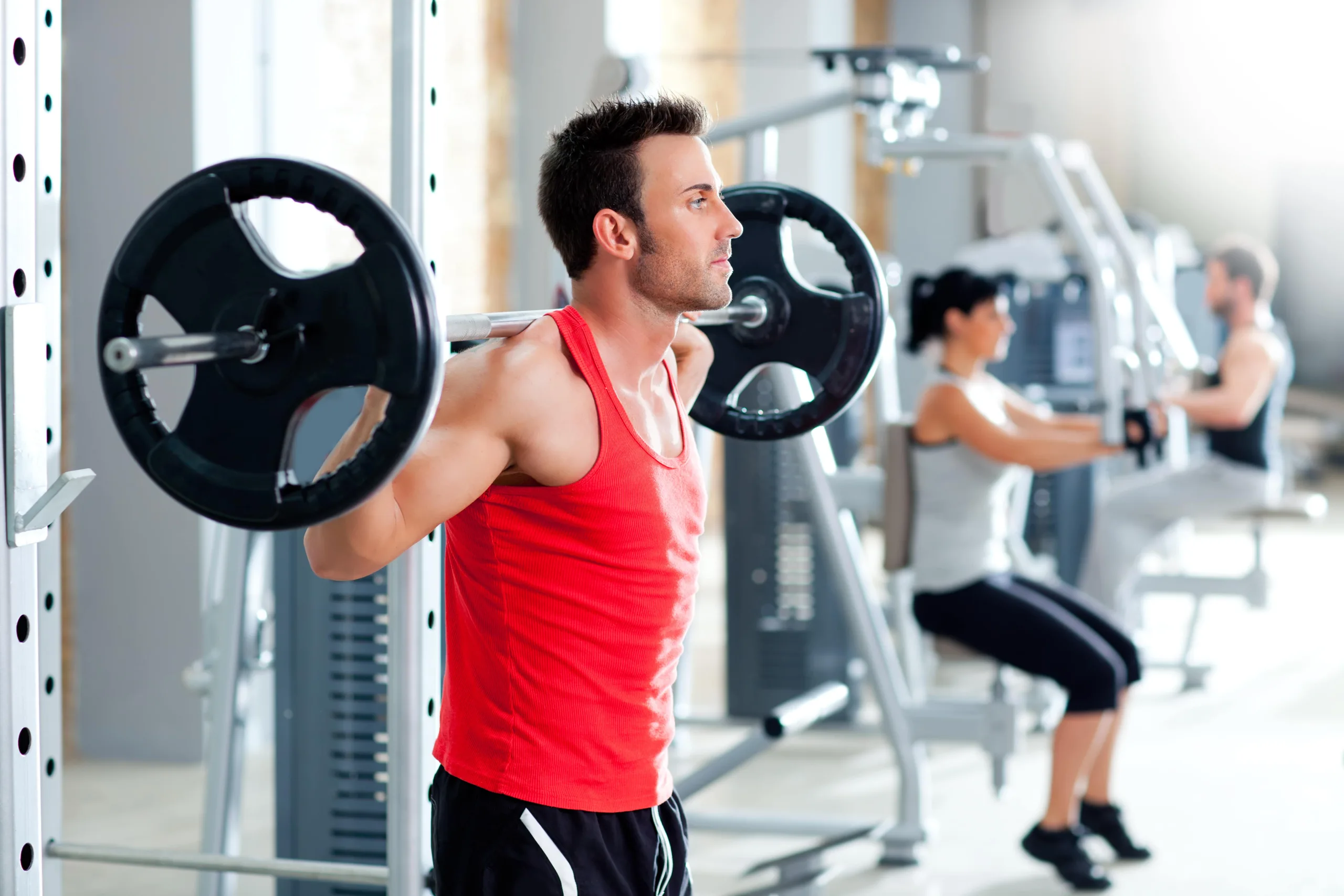 Man in weightroom wearing a red shirt and black shorts with squat bar across his back preparing to squat. In the background you can see a man and a woman on other weight machines and a new clean gym.